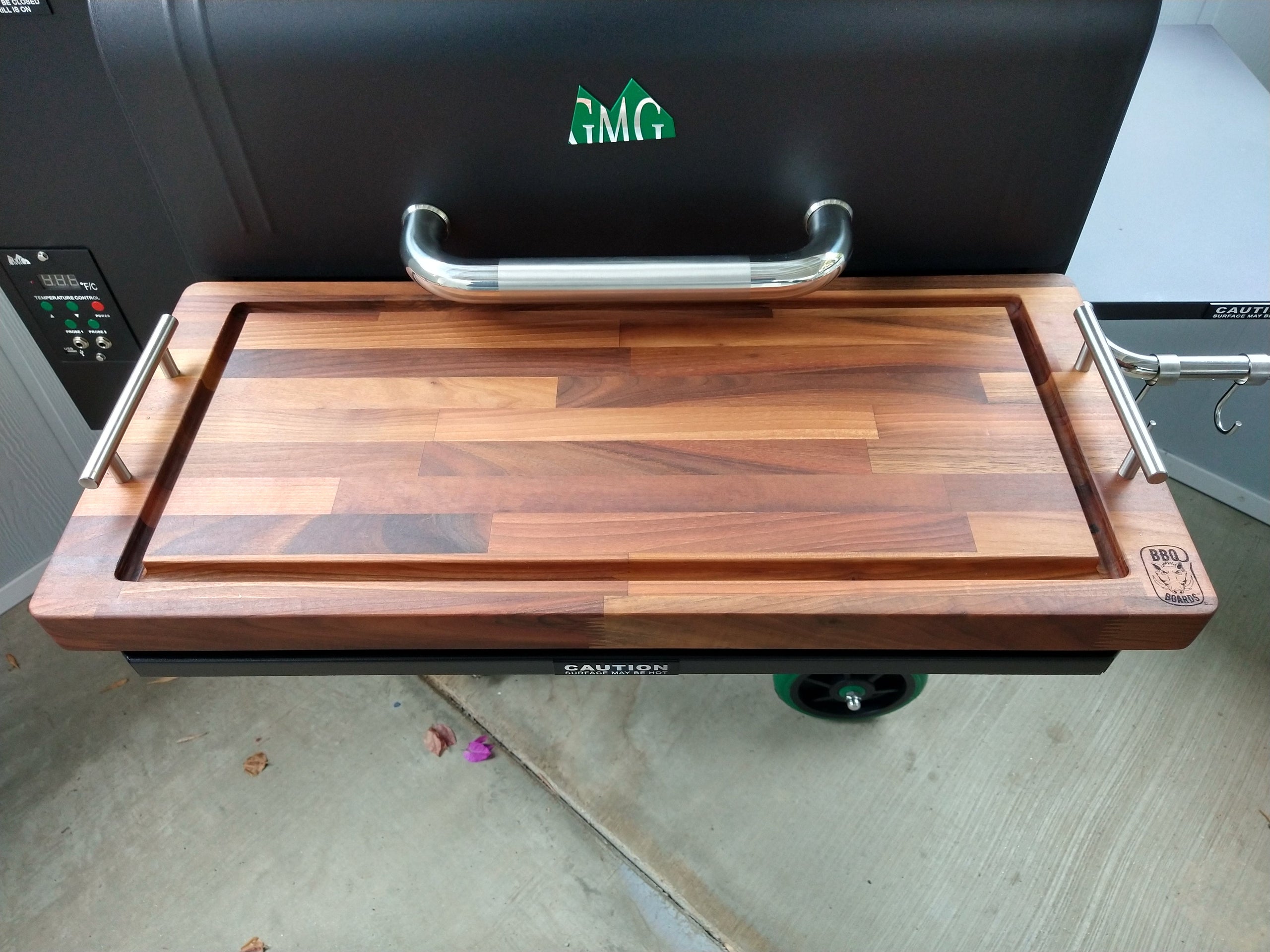 Wooden cutting board on a grill with a visible brand logo.