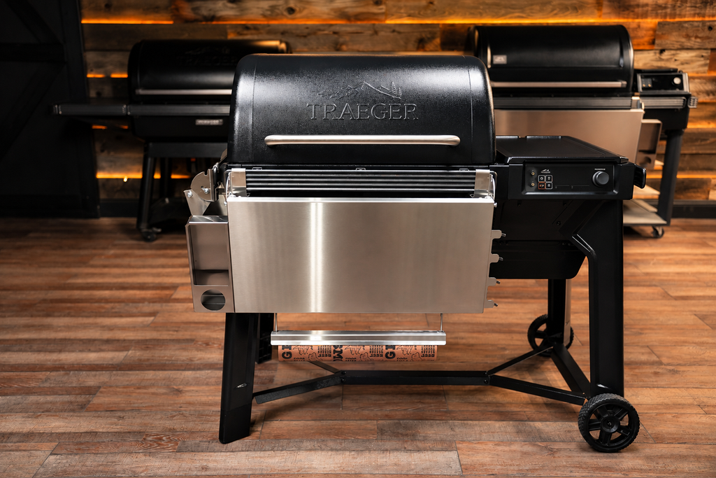 A Traeger pellet grill with a black lid and stainless steel lower drawer and legs, shown on a wood floor with other grills in the background.